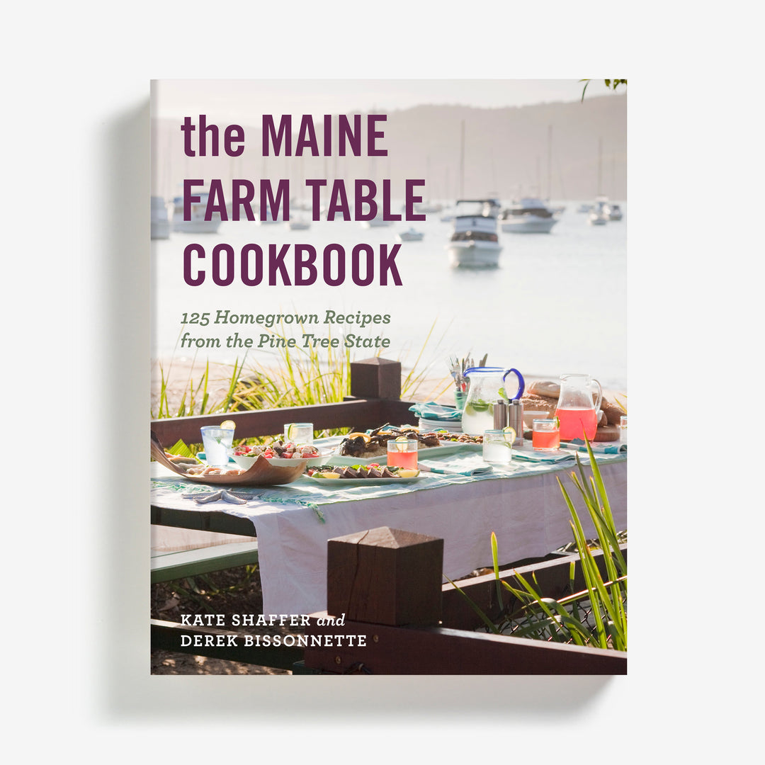 A Ragged Coast Chocolates cookbook titled 'The Maine Farm Table Cookbook' featuring local food recipes displayed in front of a scenic backdrop with a table set for dining.