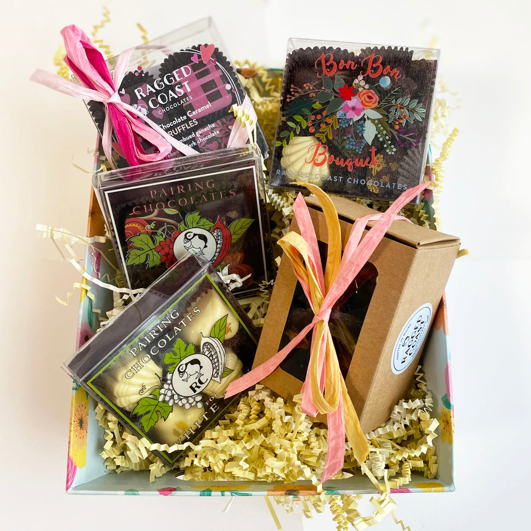 An open Mother's Day Sampler Gift Box filled with various Ragged Coast Chocolates artisan chocolates and cocoa products, adorned with pink ribbons and decorative shredded paper, perfect as a Mother's Day gift box.